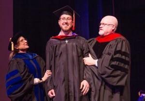 HSU graduate student at graduation standing between two professors smiling.